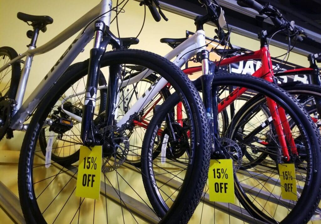 Several bicycles in silver, white, and red are displayed closely together on a rack at Jake's Bikes, each with a yellow sign on the front wheel that reads “15% OFF.”.