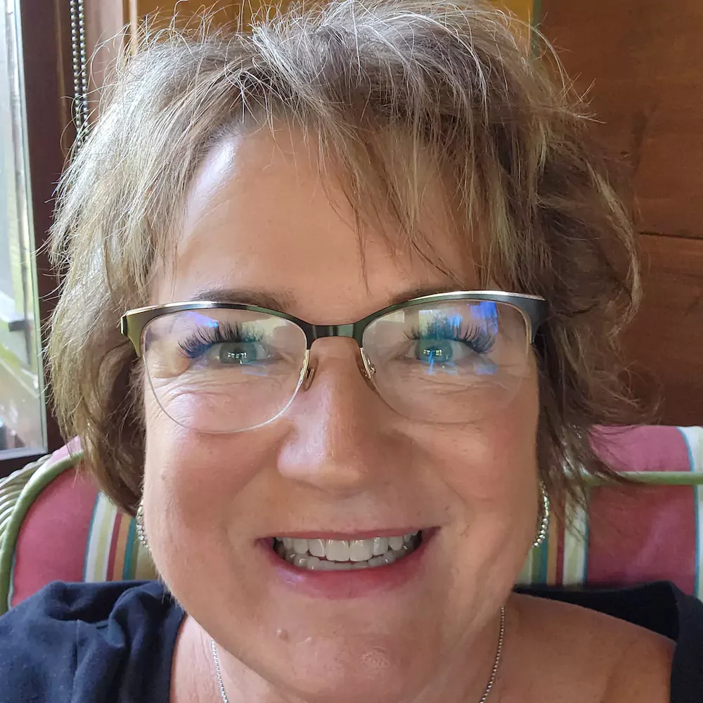 A woman with short, light brown hair and glasses smiles at the camera. She is indoors, sitting on a striped cushion with wooden walls in the background.