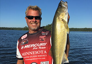 A smiling man wearing sunglasses and a red fishing jersey holds up a large fish in one hand while standing on a boat—showcasing the success of a Minnesota Fishing Guide Service—against a blue lake and tree-lined shore.