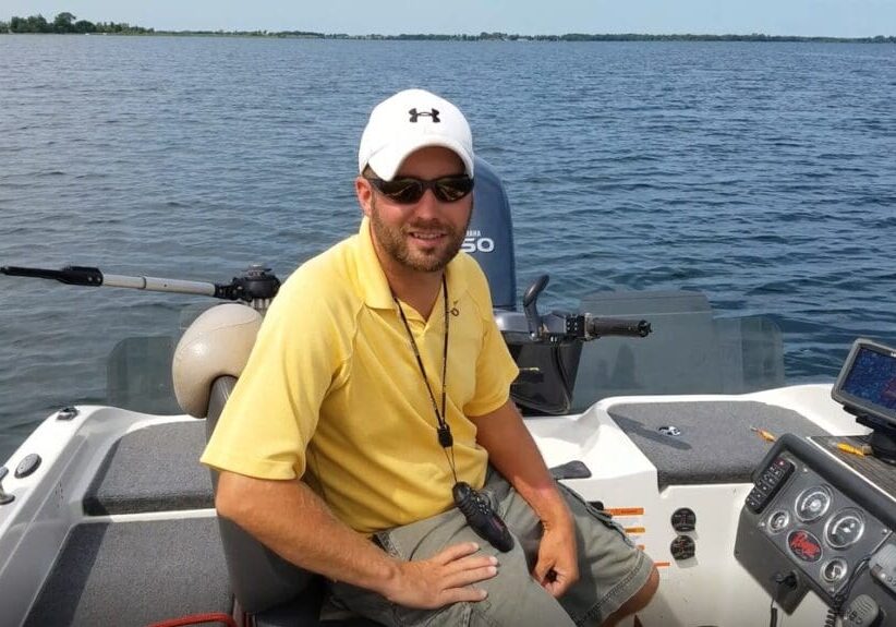 A man wearing a white cap, sunglasses, and a yellow shirt sits on a boat, smiling at the camera. Surrounded by fishing gear and boating equipment, he enjoys the day with a vast body of water and distant shoreline in the background.