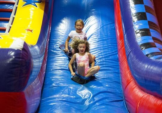Two young girls laugh and slide down a brightly colored Jump 4 Fun inflatable at the Lakes Area Indoor Park, one in a white shirt and the other in pink, surrounded by yellow, blue, and red sections with star decorations.