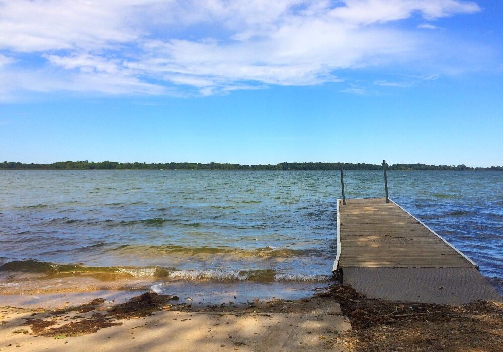 A wooden dock extends over the calm waters of Lake Andrew under a bright blue sky with scattered clouds. The shoreline features some sand and debris, with trees lining the distant horizon.