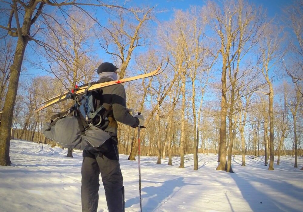A person in winter clothing stands in a snowy forest near Alexandria, holding ski poles and carrying skis and a large backpack. Bare trees surround them under a clear blue sky.