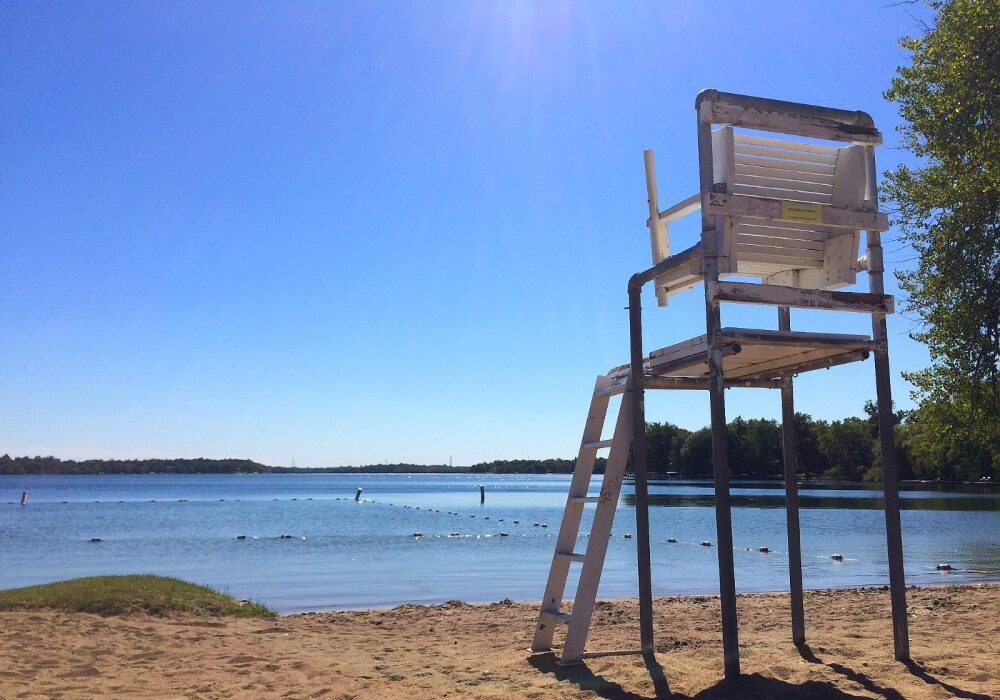 A tall, empty white lifeguard chair stands on the sandy shores of Lake Latoka by a calm lake, with trees and a clear blue sky in the background on a sunny day.