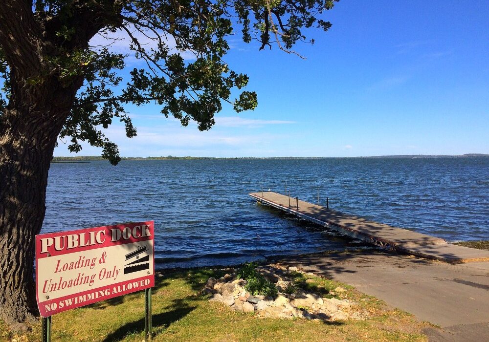 A public dock extends into the calm waters of Lake Osakis under a clear blue sky. A tree offers shade in the foreground, and a sign beside the dock reads "PUBLIC DOCK Loading & Unloading Only. No Swimming Allowed.