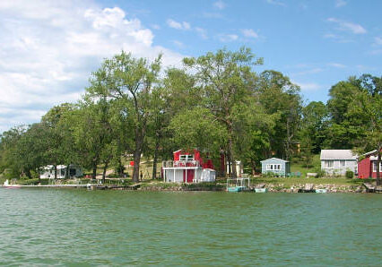 View of a lakeshore at Lake Victoria Resort, with several houses—including a prominent red one—nestled among trees and greenery under a partly cloudy sky. The calm water in the foreground enhances the peaceful setting.