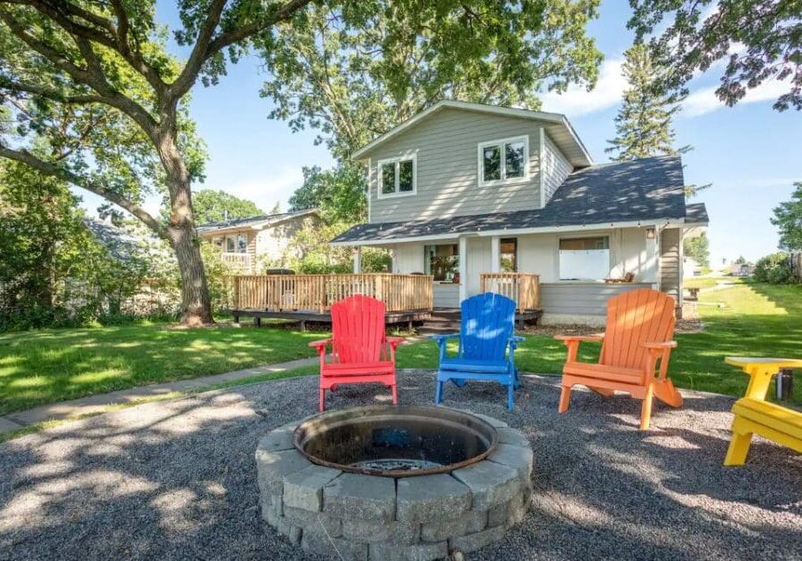 A backyard with a stone fire pit surrounded by colorful Adirondack chairs on gravel, perfect for relaxing after enjoying Lake Henry views. In the background, a two-story house with a large wooden deck is shaded by tall trees on a sunny day.