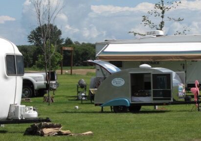 Several campers and trailers are parked on a grassy field at Lazy Days Campground under a partly cloudy sky, with lawn chairs, tables, and various outdoor items arranged beside them. Trees and vehicles can be seen in the background.