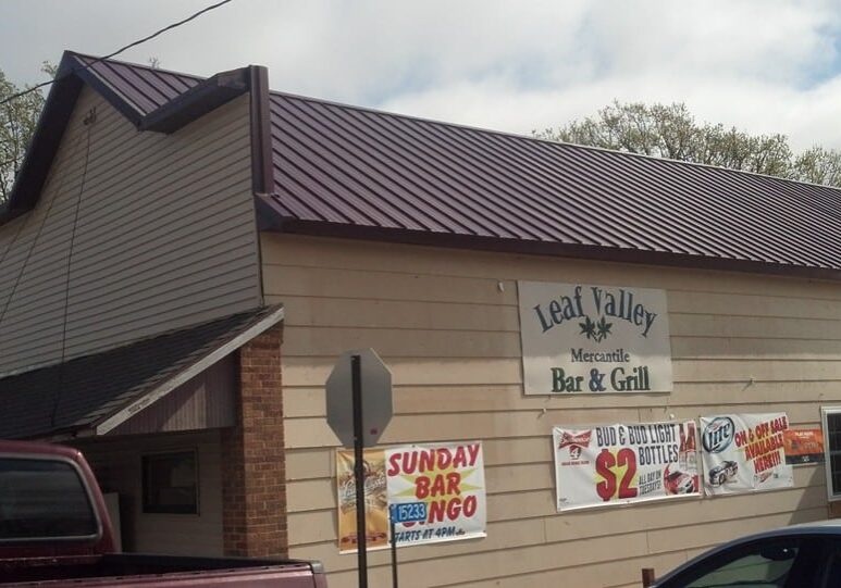 The beige building with a brown metal roof is home to Leaf Valley Mercantile Bar & Grill, where banners advertise Sunday Bar Bingo and beer specials. A maroon truck and part of a blue car are parked outside Leaf Valley Mercantile.