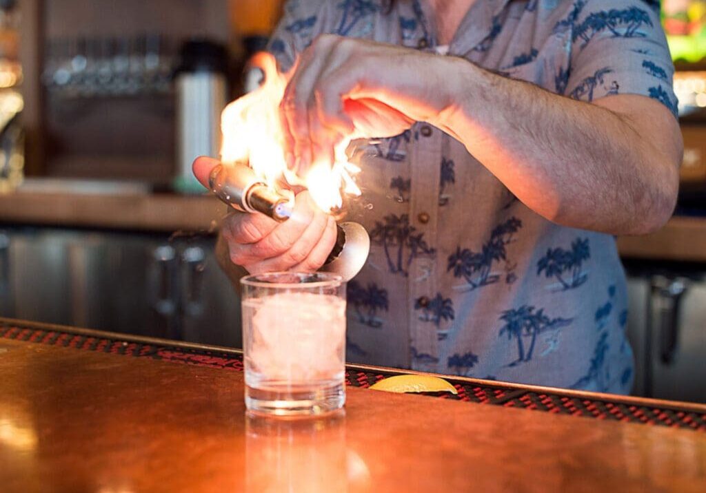 A bartender at Longtrees Woodfire Grill, in a patterned shirt, uses a torch and squeezes citrus peel, creating a burst of flame above a glass of ice on the bar counter.
