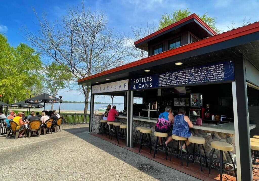 Outdoor bar, Lure Lakebar, with people sitting at the counter and nearby tables under a sunny blue sky. Trees with spring foliage surround the area, a lake is visible in the background, and live music adds to the lively atmosphere.