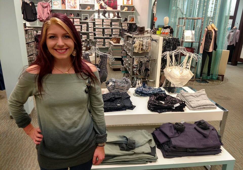 A smiling woman with long, partly reddish-blonde hair stands in a Maurices clothing store beside a display table with folded sweaters and bras. Shelves with shoes and more clothing are visible in the background.