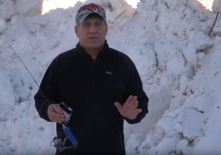 A man wearing a black jacket and a baseball cap stands in front of a large snowbank, holding a fishing rod in one hand and gesturing with the other, resembling angler Mike Frisch.