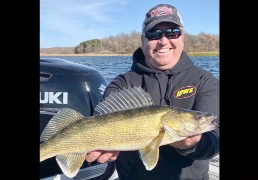 A smiling person wearing sunglasses and a black hoodie holds a large walleye fish on a boat, with a Suzuki motor and a lake in the background—just another great day of fishing with Mike Frisch.