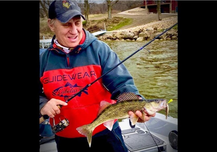 A man in a red and navy jacket, possibly Mike Frisch, holds a fishing rod and proudly displays a freshly caught fish while standing on a boat in a lake near a rocky shoreline.