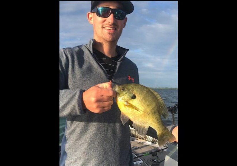 Mike Frisch, wearing sunglasses, a cap, and a gray jacket, smiles while holding a yellowish fish on a boat, with water and a partly cloudy sky in the background.