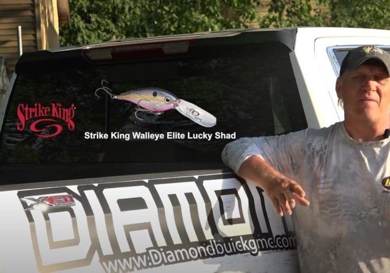 A man in a cap and camo shirt stands by a white truck with "DIAMOND" and a web address on the tailgate. The rear window displays a Strike King fishing lure ad, a large fishing lure image, and the name Mike Frisch.