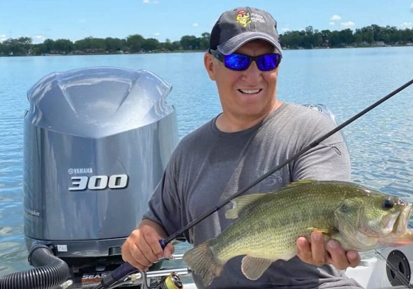 A man wearing sunglasses and a cap smiles while fishing, holding a large fish on a boat in a lake, with a Yamaha 300 outboard motor behind him and trees in the background.