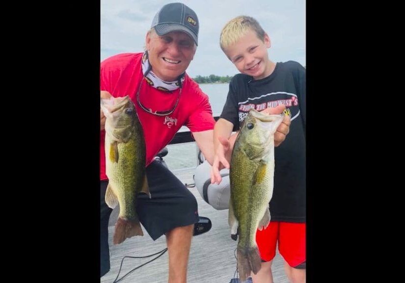 An adult and a smiling boy stand on a dock holding large fish they caught while fishing, with a lake and cloudy sky in the background. Both appear happy and proud of their catches.