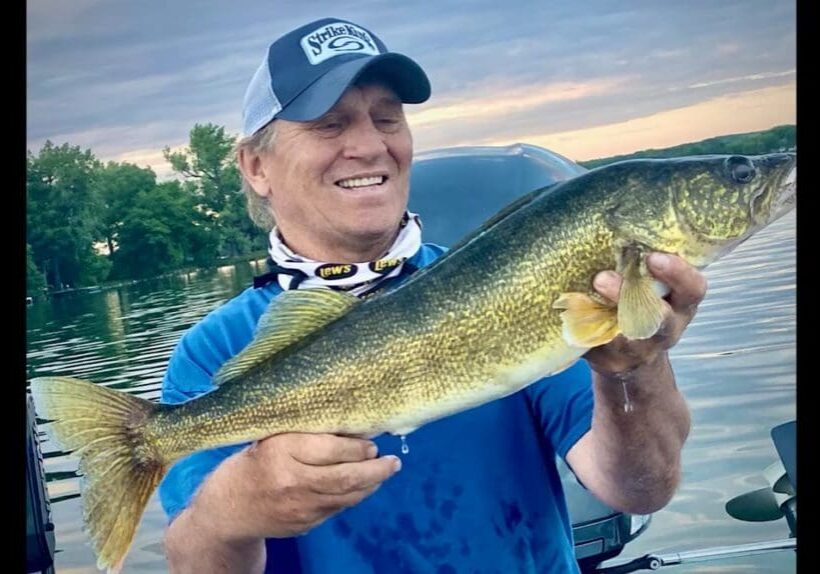 A smiling person in a blue shirt and cap shows off their fishing skills, holding a large fish with greenish-yellow scales on a boat, with water and trees in the background during sunset.