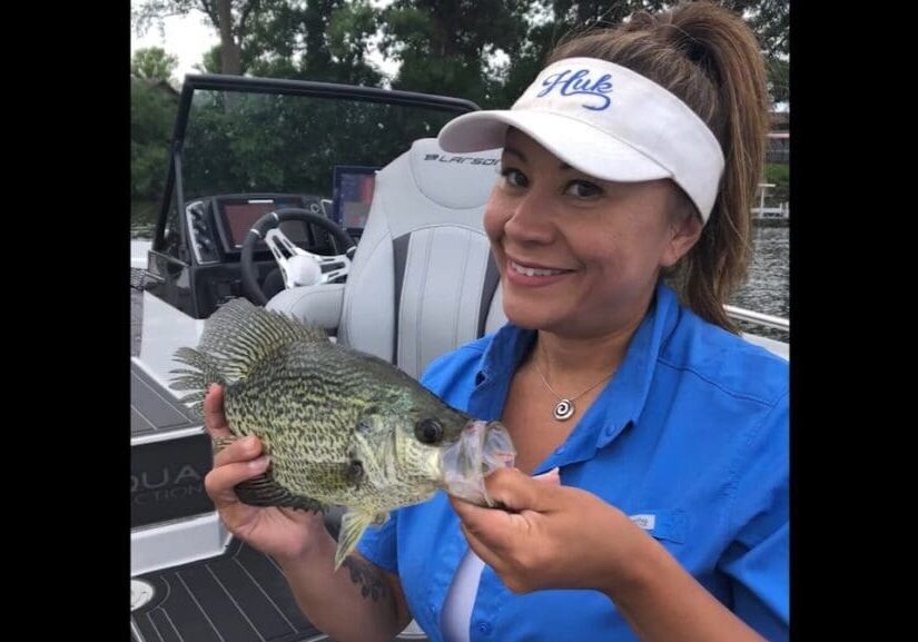 A smiling woman in a blue shirt and white visor holds a large fish beside a boat on the water, with trees in the background—just like an angler from Mike Frisch’s fishing adventures.