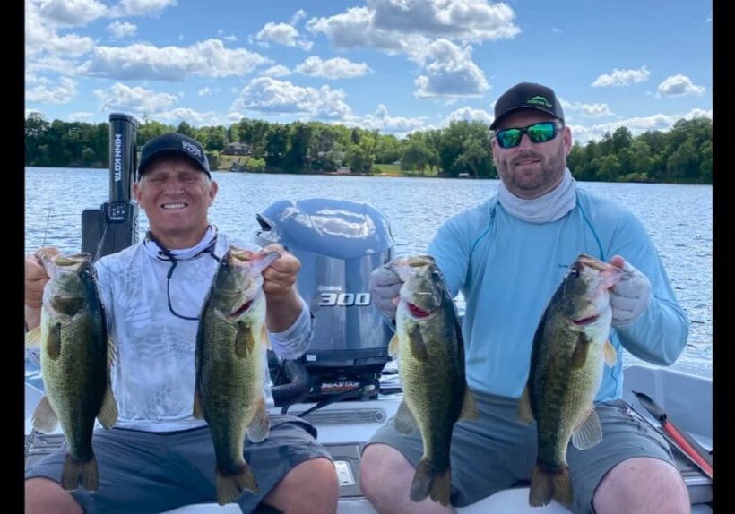 Two men, including Mike Frisch, sit on a boat holding up large bass they caught, smiling at the camera. They are on a lake surrounded by trees and set against a blue sky with scattered clouds.