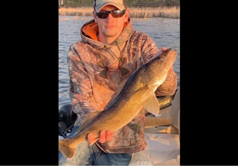 A person wearing a camouflage hoodie, sunglasses, and a cap holds a large walleye fish while fishing from a boat on a lake, with reeds and trees visible in the background.