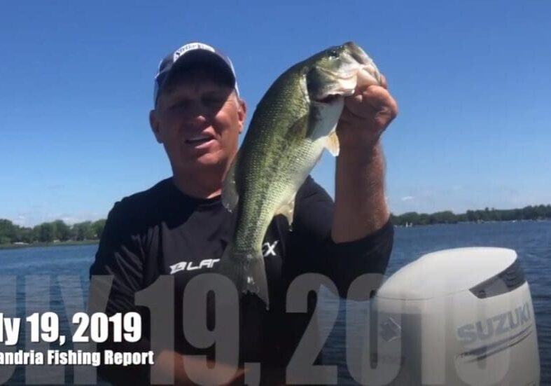 A man on a boat holds up a large fish with a lake and trees in the background. Text on the image reads "July 19, 2019 Alexandria Fishing Report with Mike Frisch." A Suzuki boat motor is visible on the right.