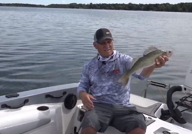 A man, possibly Mike Frisch, sits in a boat on a lake holding up his catch with a proud smile. He sports a cap, blue camouflage shirt, shorts, and a neck gaiter. Trees line the distant shore beneath a cloudy sky.