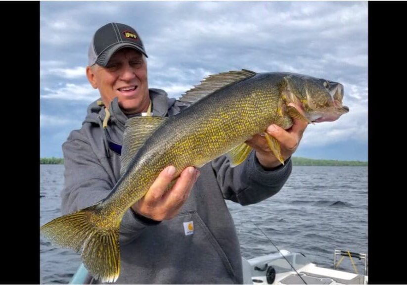 A person in a gray hoodie and cap, possibly Mike Frisch, smiles while holding a large yellow-green fish with sharp fins on a boat, with a lake and cloudy sky in the background.