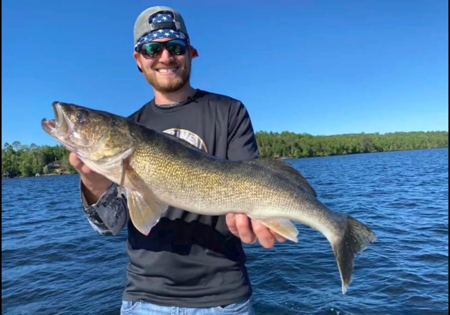 A smiling man wearing sunglasses and a cap shows off his catch while fishing on a boat in a lake, with trees and a clear blue sky in the background.