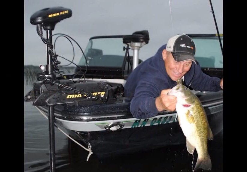 A man in a cap and blue jacket leans out of a fishing boat to hold up a large fish he has caught, smiling proudly as he displays his fishing success over the water.