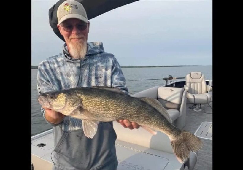 A man wearing glasses, a cap, and a blue hoodie stands on a boat holding a large fish with both hands. The water and sky are visible in the background, capturing the excitement of Mike Frisch's fishing adventure.