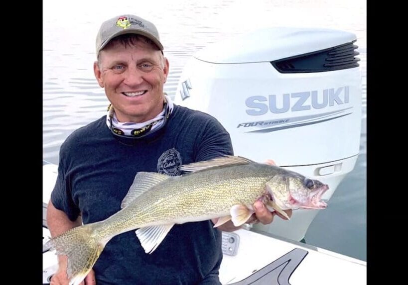 A smiling man in a cap and t-shirt proudly holds a large fish on a boat during fall fishing, with water and a Suzuki outboard motor visible in the background.