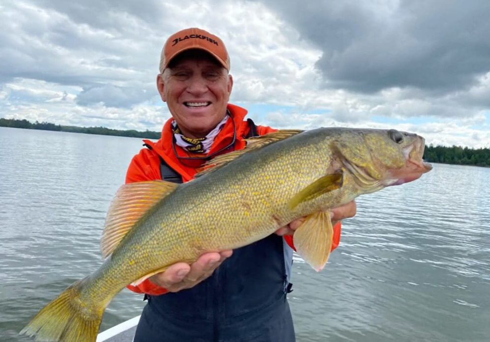 Smiling man in an orange cap and jacket, possibly Mike Frisch, holds a large walleye with yellowish scales while standing on a boat in a lake, with trees and cloudy sky in the background.