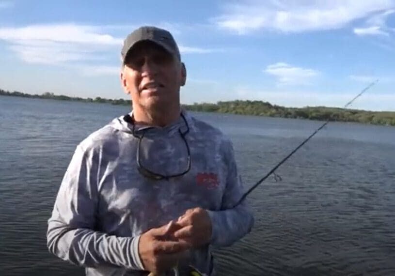 A man in a cap and sunglasses rests a fishing rod while standing by a large lake under a blue sky with scattered clouds, hoping to catch crappies. Trees and shoreline are visible in the distance.