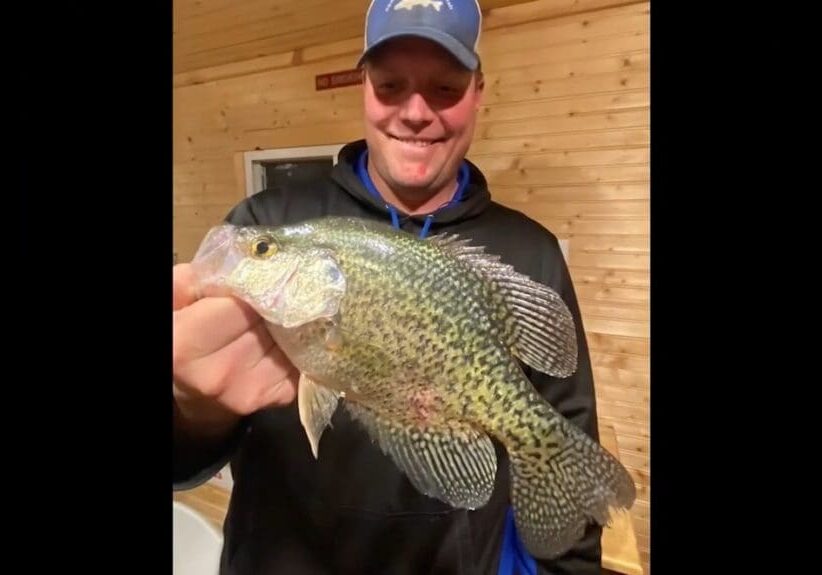 A smiling man in a blue cap and black hoodie, identified as Mike Frisch, holds a large, spotted crappie fish indoors against a wooden wall background.