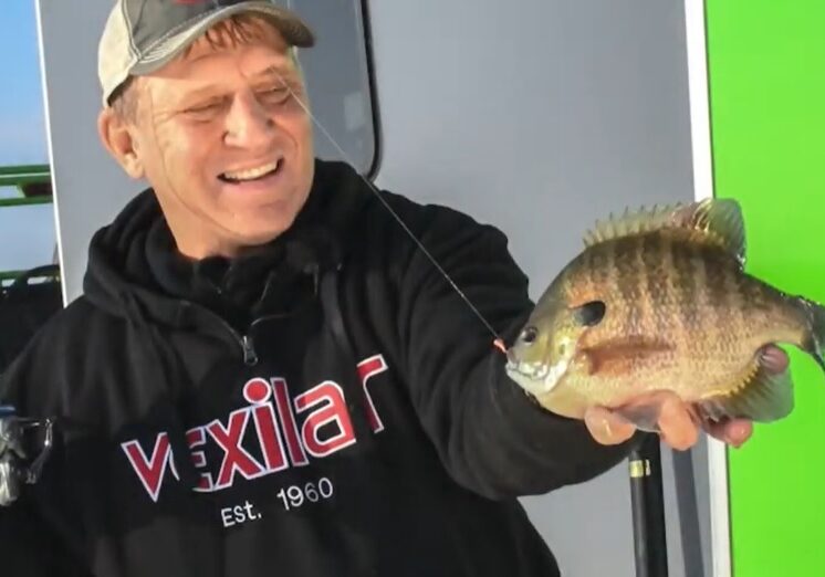 A smiling man, Mike Frisch, in a black "Vexilar" hoodie holds up a freshly caught fish, displaying it proudly. The background includes a green and white wall.