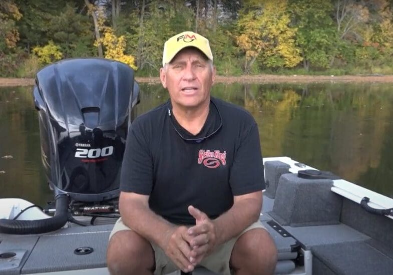A man wearing a yellow hat and black shirt sits on a fishing boat in front of a lake, surrounded by autumn-colored trees. He faces the camera and appears to be speaking about his fishing experience.