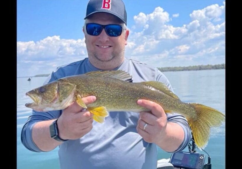 A smiling man, possibly Mike Frisch, wearing sunglasses and a baseball cap holds a large fish while standing on a boat, with calm water and a partly cloudy sky in the background.