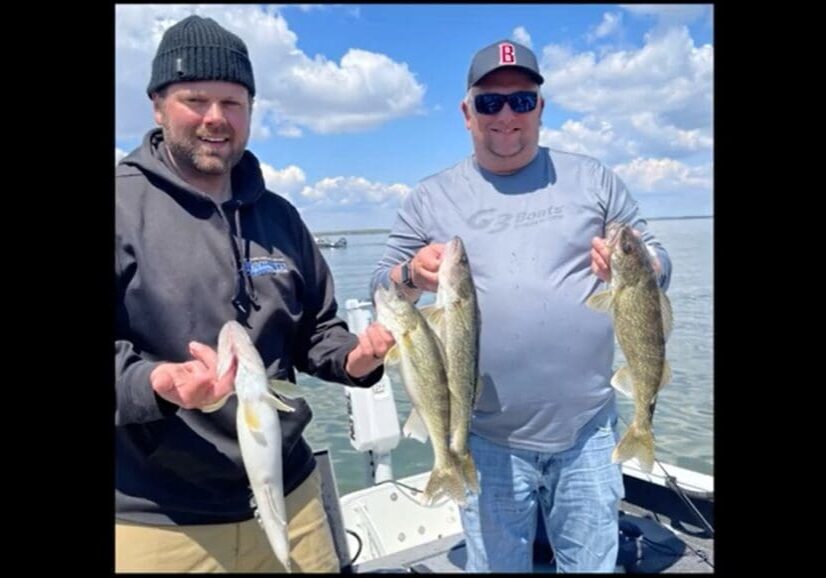 Two men stand on a boat, proudly holding up several large fish they caught while fishing, smiling at the camera. The sky is partly cloudy, and calm water stretches out behind them. Both are dressed in casual outdoor clothing.