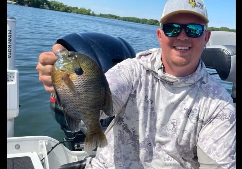 A smiling person wearing sunglasses and a hoodie—possibly named Mike—holds up a large fish while sitting in a boat on a lake, with water and trees visible in the background.