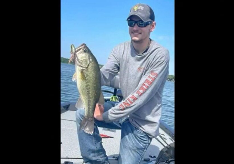 A man in sunglasses and a blue cap smiles while proudly holding his catch—a large fish—on a boat, with water and sky in the background.