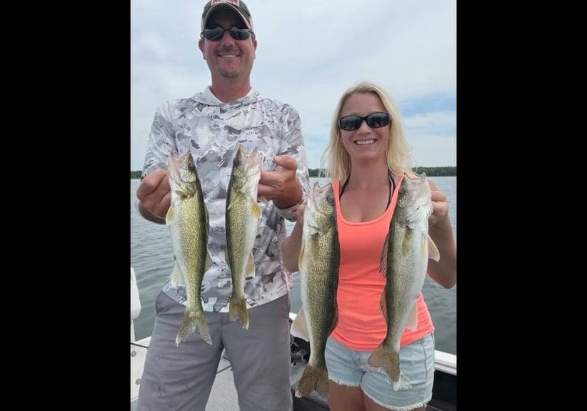 A man and a woman on a boat each hold up two walleye, smiling at the camera. The man wears a camo shirt, cap, and sunglasses; the woman sports sunglasses, a neon tank top, and shorts. Water and sky stretch into the background.