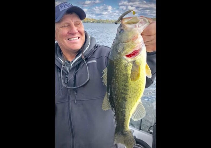 A smiling person wearing a cap and jacket holds up a large bass fish with one hand on a fishing boat, water and a cloudy sky in the background.