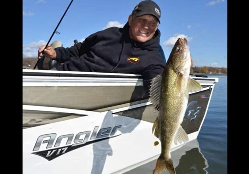 A person smiling and holding a fishing rod sits in a boat labeled "Angler V17" on Alexandria’s sparkling waters, proudly holding up a large fish above the water on a clear day.