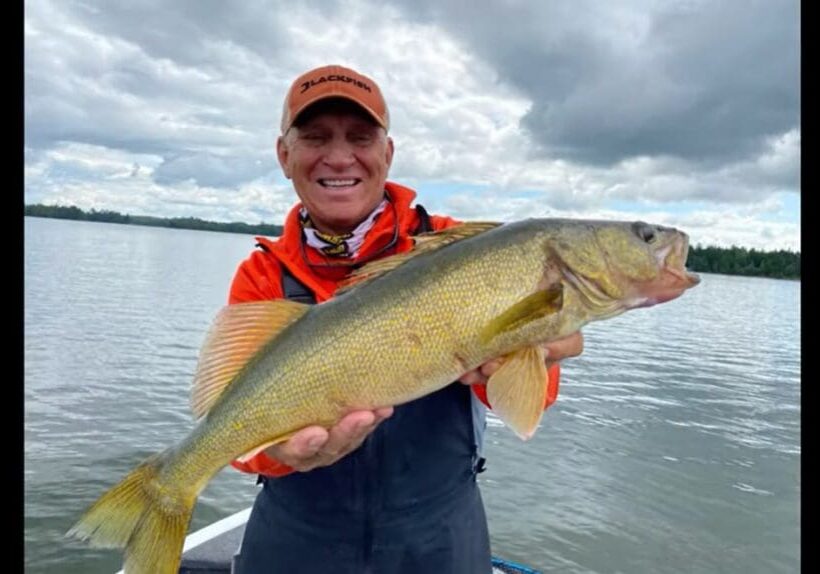 A smiling person in an orange cap and jacket holds a large yellow fish while standing on a boat, enjoying fall fishing on a lake with cloudy skies and distant trees in the background.