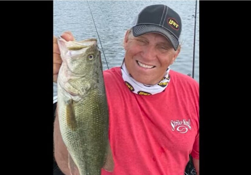 Mike, a smiling man in a red shirt and black cap, holds up a large fish in front of a lake, with fishing gear visible in the background.