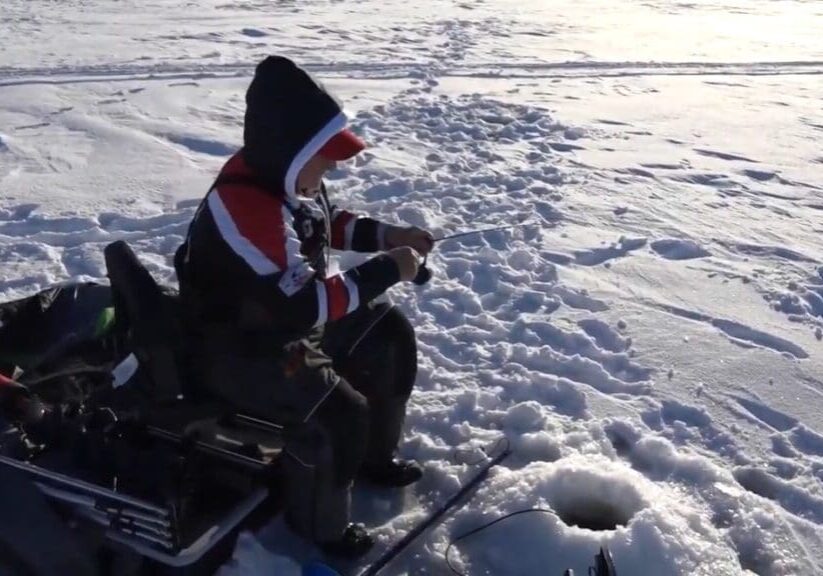 A person sits on a folding chair ice fishing, dressed in winter clothing with a hood, much like mike frisch. They hold a fishing rod over a hole in the snow-covered ice, surrounded by equipment and footprints.