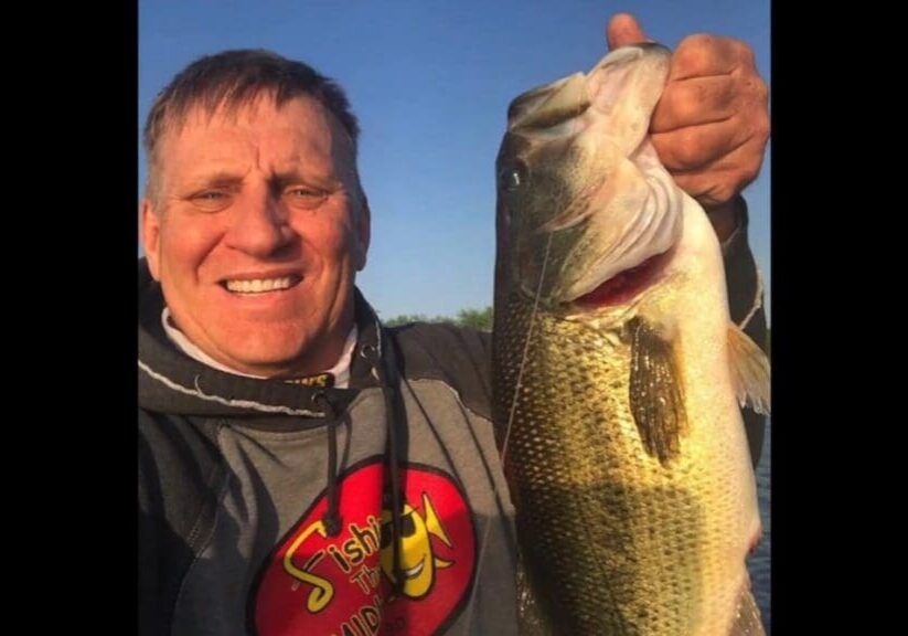 A smiling man in a gray hoodie with a red and yellow “Fishing” logo holds up a large fish under a clear blue sky. Mike Frisch proudly shows off his catch, capturing the perfect fishing moment.